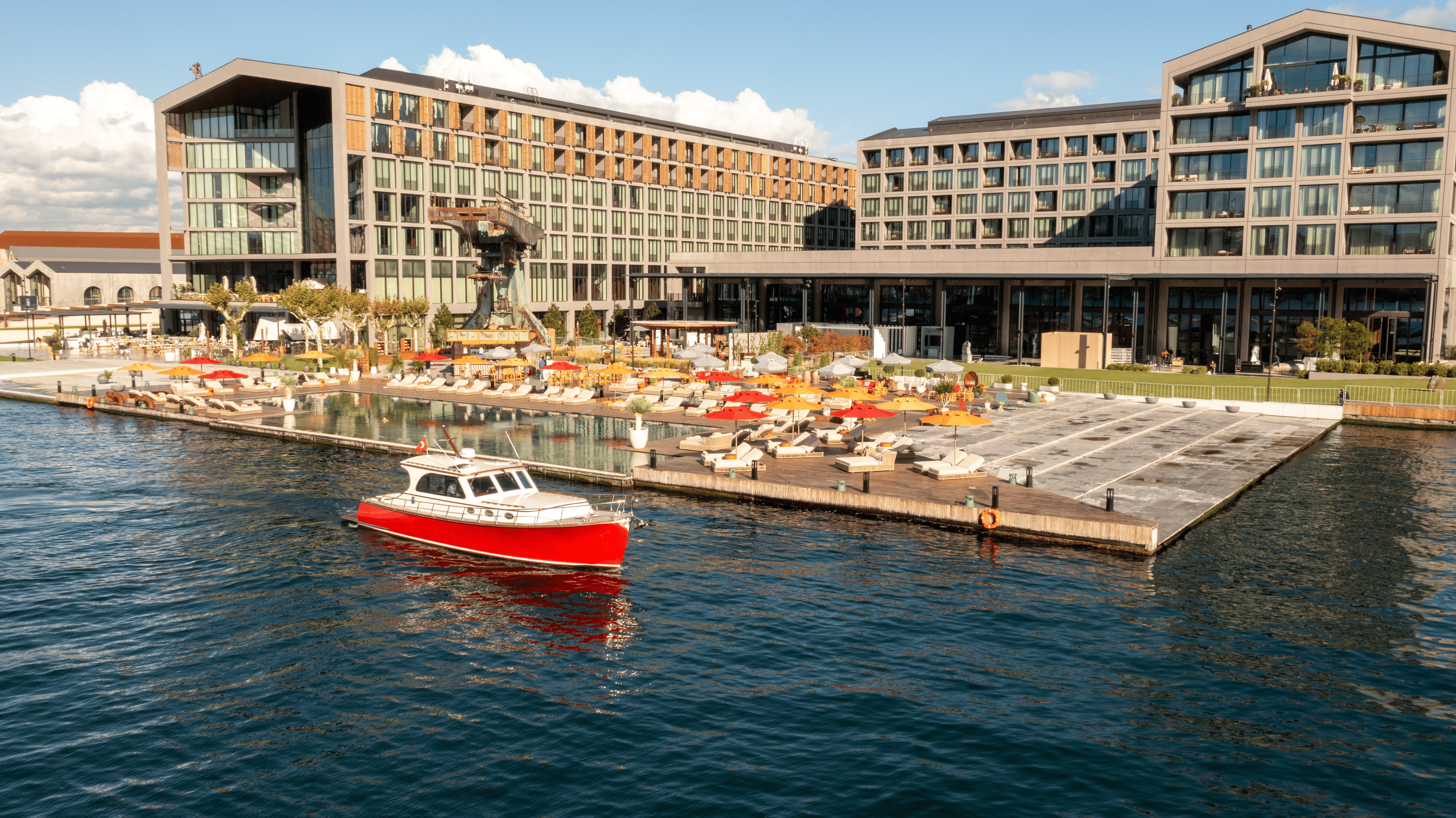 Rixos Tersane Стамбул қонақ үй кешенінің кең кадры, шезлонгтары мен қызыл және қызғылт сары түсті қолшатырлары бар жағалаудағы палубаға қарайтын екі үлкен заманауи ғимараты бар. Алдыңғы қатардағы көк суда ақ-қызыл шағын қайық жүзіп келеді.