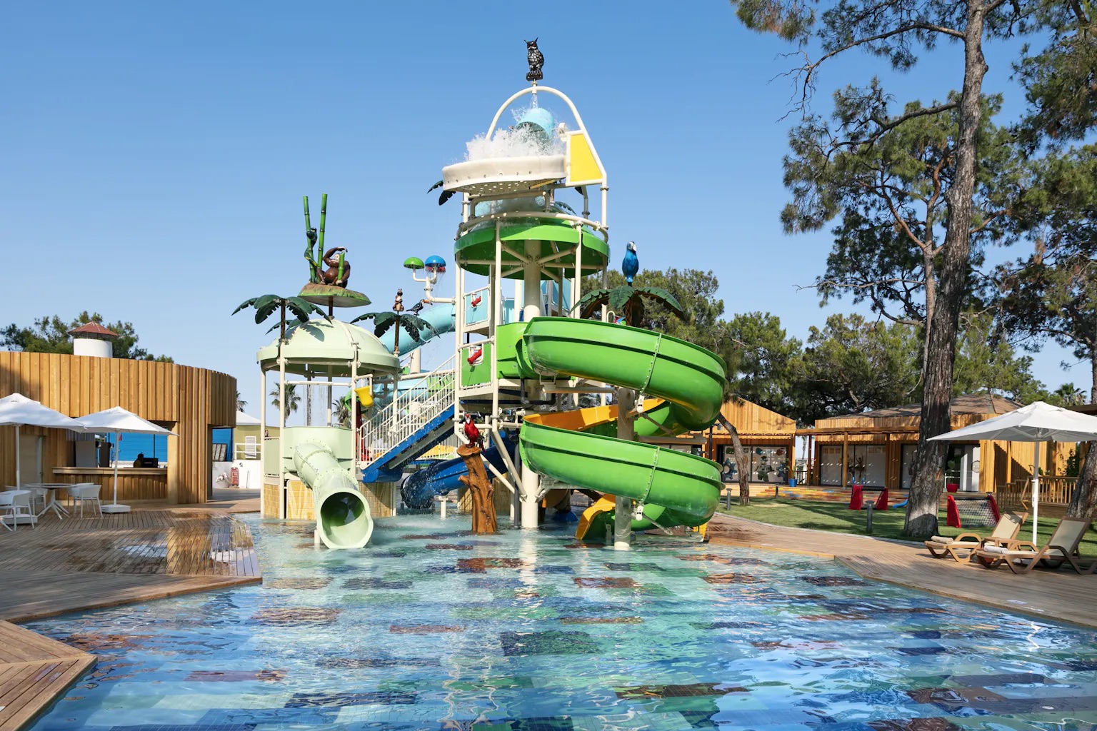 Buitenwaterpretpark met meerdere groene buisglijbanen, dierenversieringen, palmbomen en een grote emmer die water morst, gelegen in een zonnige omgeving met bomen en houten gebouwen.