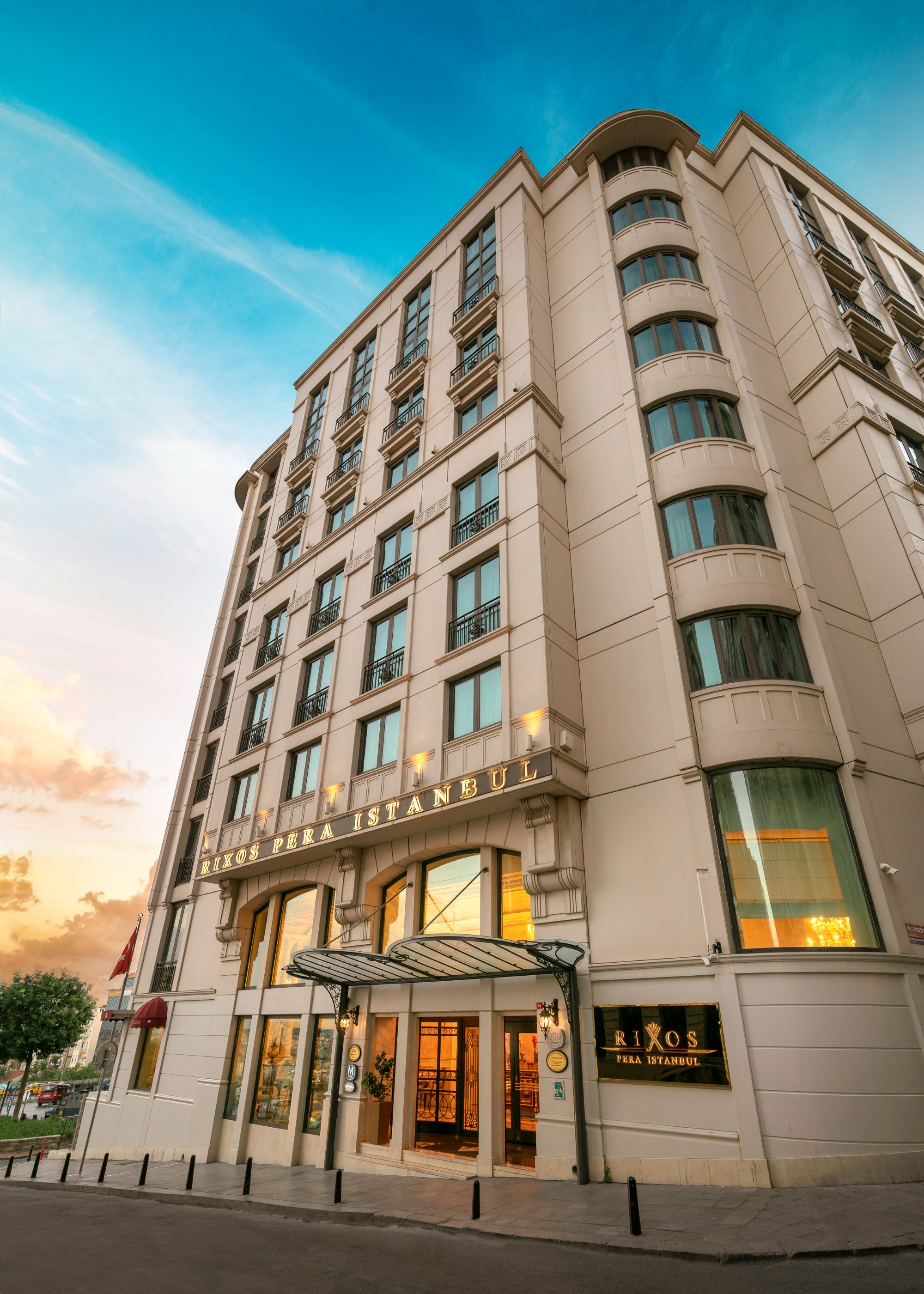 O fotografie dramatică, cu unghi ascendent, a exteriorului hotelului Rixos Pera Istanbul. Clădirea înaltă, tradițională, din piatră, are mai multe rânduri de ferestre și o intrare principală acoperită în partea de jos, toate pe fundalul unui cer albastru strălucitor și înnorat.