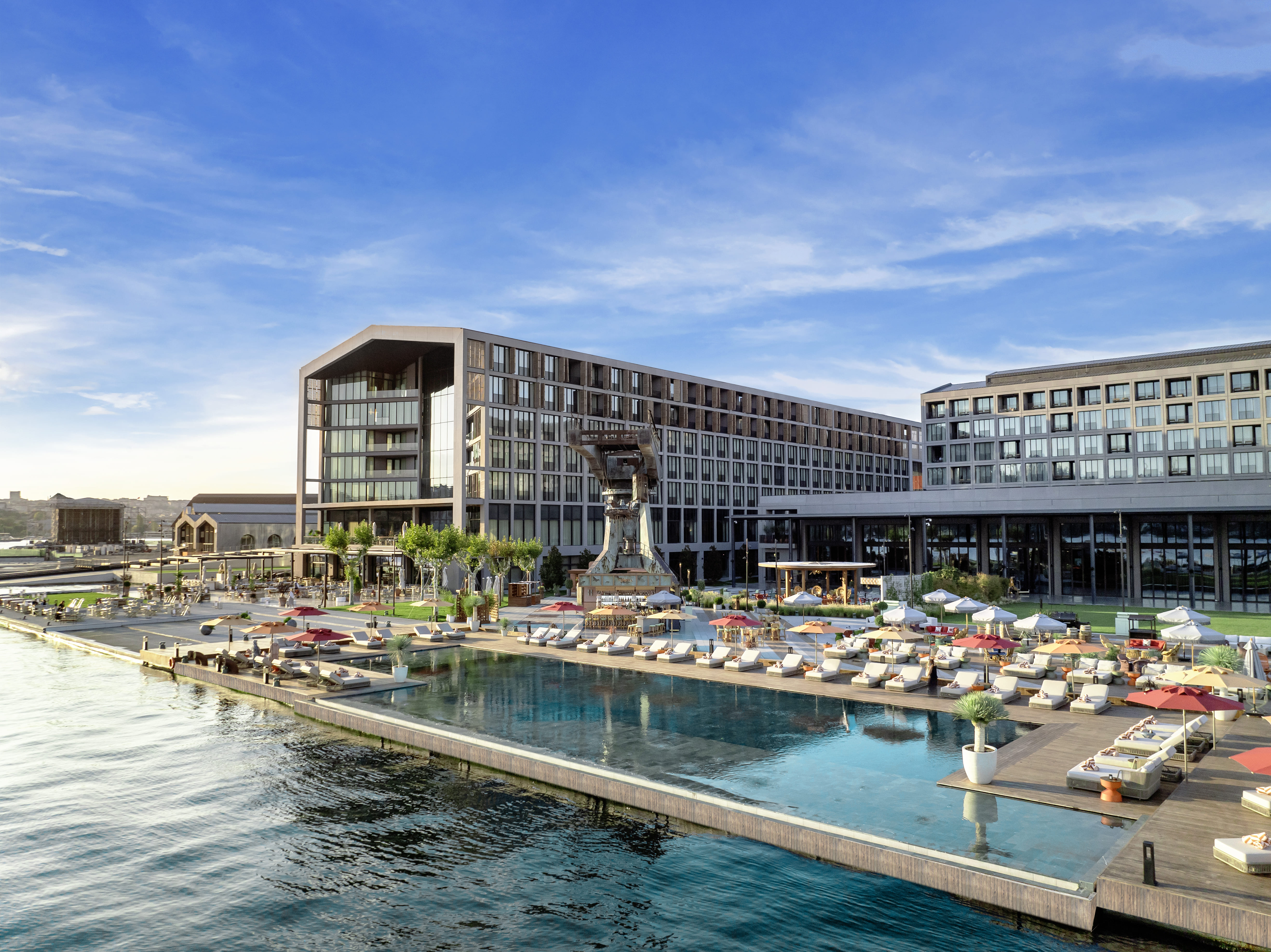 Um moderno hotel à beira-mar, o Rixos Tersane Istanbul apresenta grandes janelas de vidro, uma piscina exterior de beiral infinito, espreguiçadeiras, guarda-sóis e jardins paisagísticos com vista para águas calmas sob um céu azul claro.
