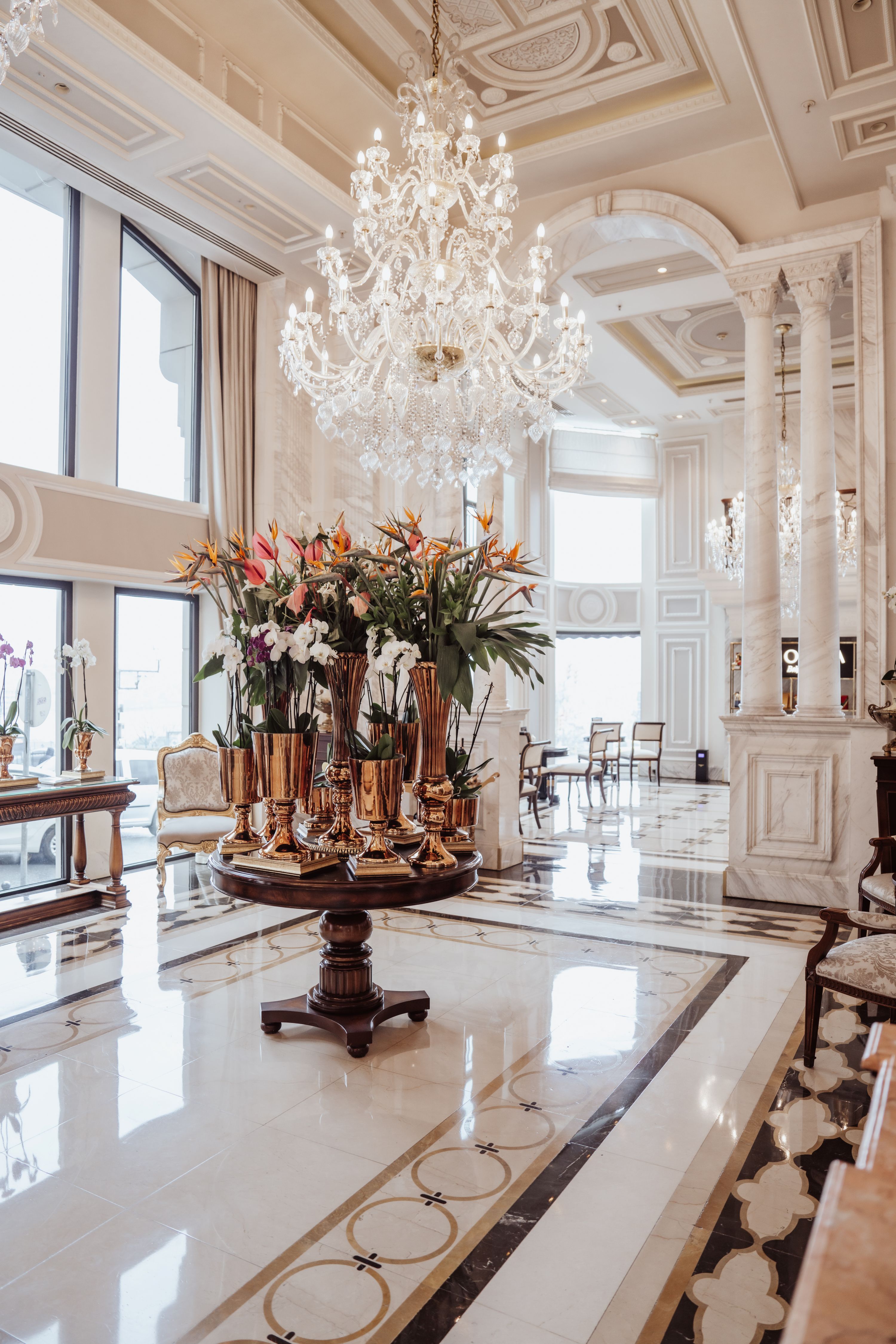 Розкішне, світле фойє готелю Rixos Pera Istanbul з високими стелями, великими вікнами, великою люстрою та центральним круглим столом, прикрашеним високою квітковою композицією. Декор вирізняється елегантними біло-золотими акцентами.